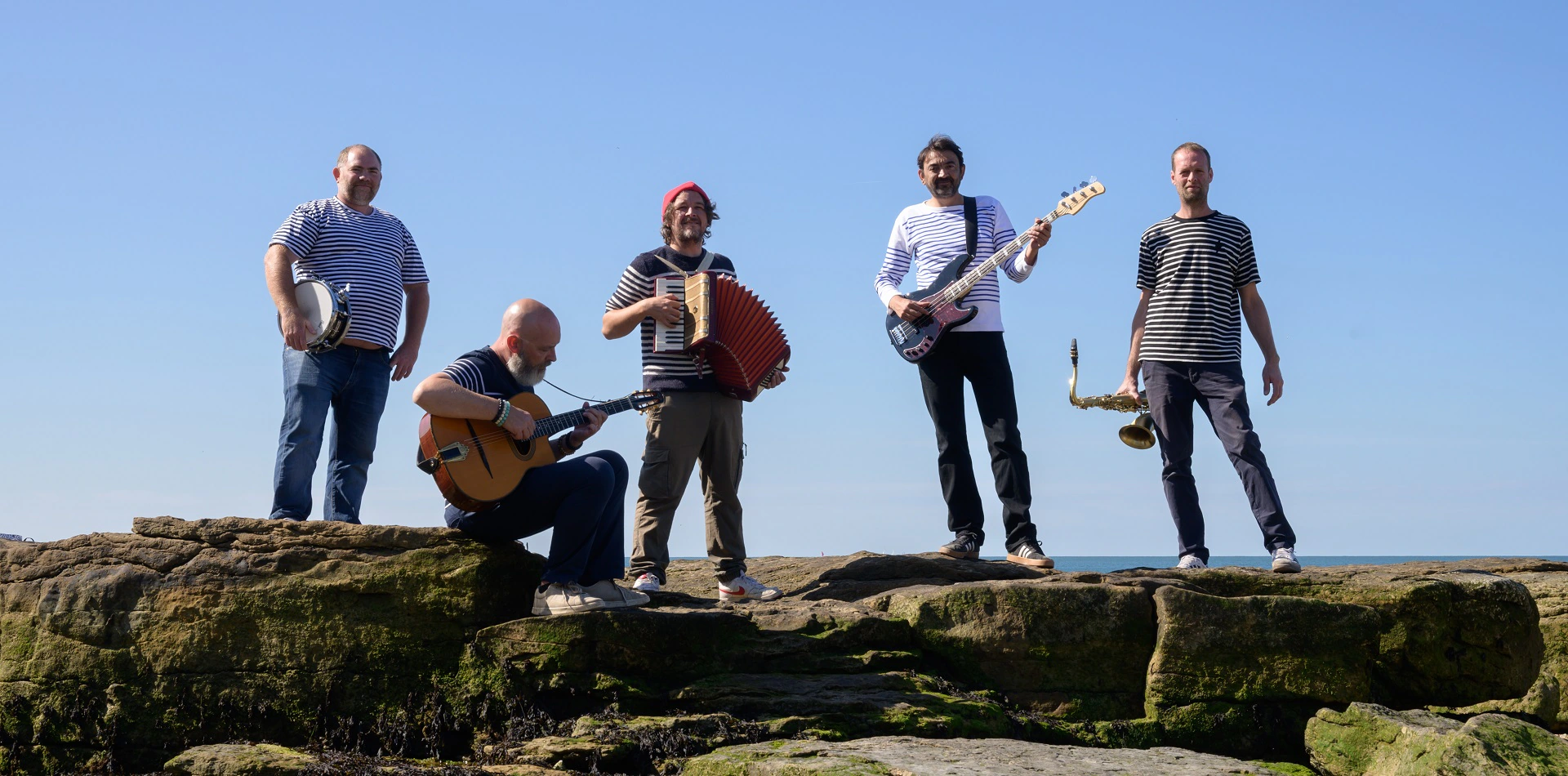 Les cinq musiciens de Lily en marinière, instruments à la main, sur les rochers face à la mer
